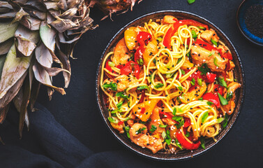 Stir fryed egg noodles with chicken, pineapple, paprika, green onion, soy sauce and sesame seeds in bowl. Asian cuisine dish. Black table background, top view