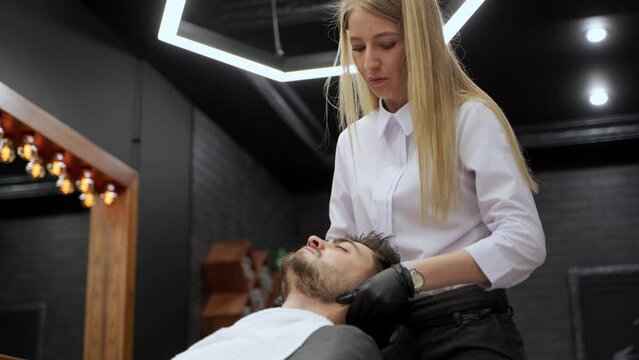 Woman performs precise face massage on man in modern barbershop. Professional beautician at work in mens spa salon. Relaxing facial treatment, skincare routine, male grooming, wellness concept.
