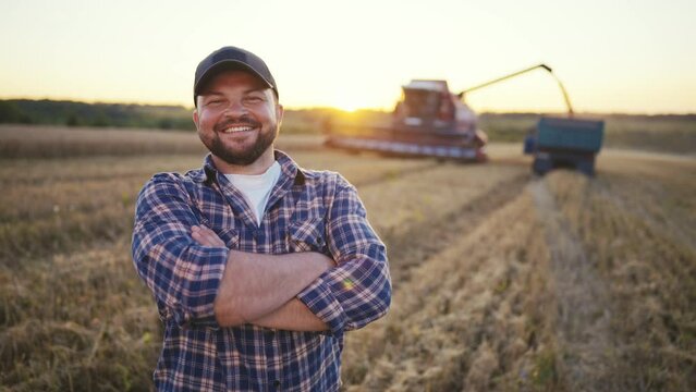 Happy bearded farmer in cap looking at camera. Farmer on agricultural wheat field with combine harvester and dump truck on background. Harvesting, farming at sunset. Food production, agribusiness.