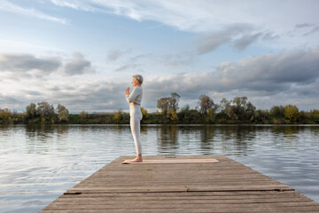 Fototapeta na wymiar An adult woman in a white suit performs yoga asanas on a pier by the river. Tadasana. Mountain Pose