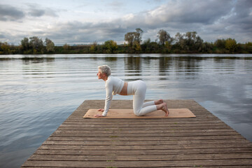 Fototapeta na wymiar An adult woman in a white suit performs yoga asanas on a pier by the river. Majariasana . Cat pose, bending down