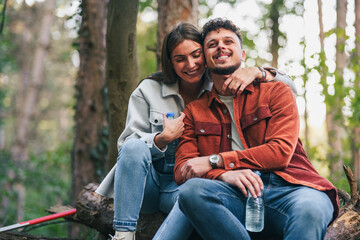 A girl is hugging her boyfriend with a smile while sitting on a branch after a long hike. They are...