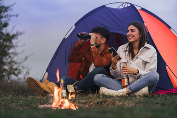 A couple is enjoying themselves in front of a tent by a campfire, the boy is holding binoculars in...