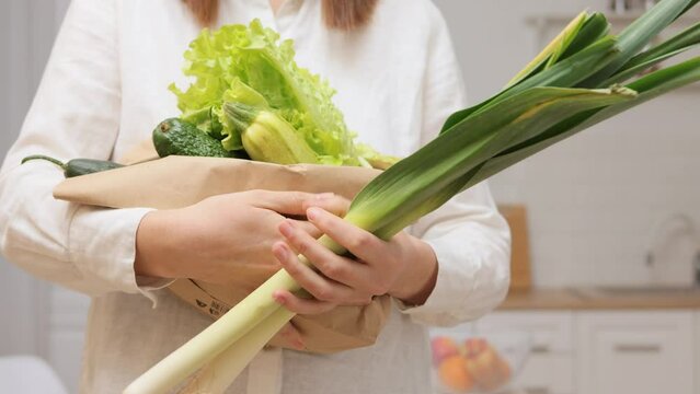 Unknown woman wearing white shirt holding in hands paper bag with vegetables showing leek for preparing salad and other healthy meal in kitchen at home