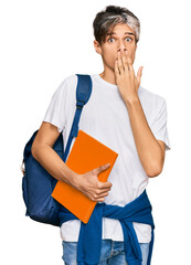 Young hispanic man wearing student backpack and holding books covering mouth with hand, shocked and afraid for mistake. surprised expression