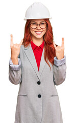 Young redhead woman wearing architect hardhat shouting with crazy expression doing rock symbol with hands up. music star. heavy concept.