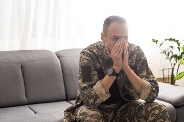 Thoughtful military man staring aside, holding palms by mouth, sitting on couch at home. Young soldier visiting psychologist, suffering from posttraumatic stress, closeup photo, copy space - obrazy, fototapety, plakaty