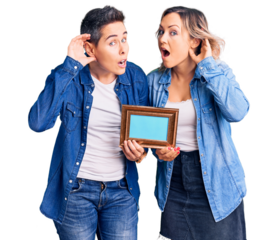 Couple of women holding empty frame smiling with hand over ear listening an hearing to rumor or gossip. deafness concept.