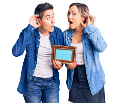 Couple of women holding empty frame smiling with hand over ear listening an hearing to rumor or gossip. deafness concept.