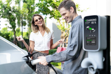 Young couple travel with EV electric car charging in green sustainable city outdoor garden in...