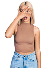 Beautiful young blonde woman wearing casual style with sleeveless shirt peeking in shock covering face and eyes with hand, looking through fingers with embarrassed expression.