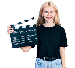 Young caucasian woman holding video film clapboard looking positive and happy standing and smiling...