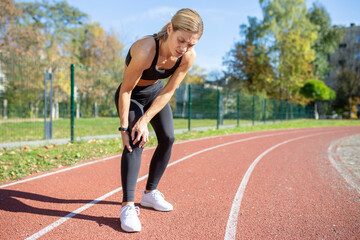 Female athlete in sportswear pausing her exercise due to a painful knee injury on a sunny outdoor...