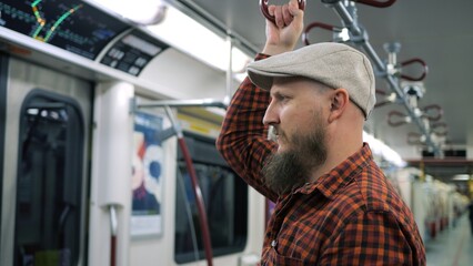 Thoughtful man in a cap holds handrail in subway. Close-up of bearded man in red checkered shirt goes on subway to downtown. Concept gloomy atmosphere in subway makes you stuck in your thoughts