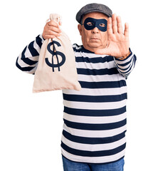Senior handsome man wearing burglar mask holding money bag with open hand doing stop sign with...