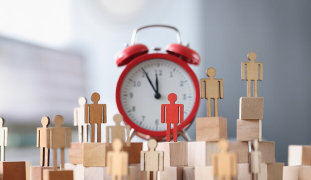 Wooden figurines with one red figure stand next to alarm clock. Time management and individual approach business planning concept
