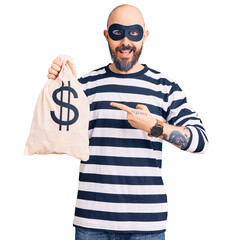 Young handsome man wearing burglar mask holding money bag with dollar symbol smiling happy pointing...