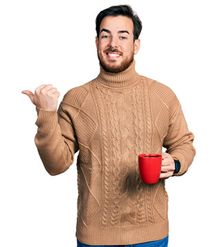 Young hispanic man holding coffee pointing thumb up to the side smiling happy with open mouth