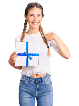 Beautiful caucasian woman with blonde hair holding gift pointing finger to one self smiling happy and proud
