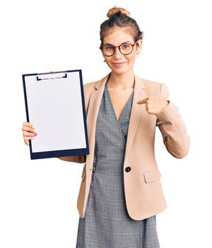 Beautiful caucasian woman with blonde hair holding clipboard with blank space pointing finger to one self smiling happy and proud