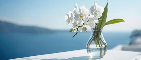 Tuinposter White orchid flower in a glass vase on a white tabletop against the backdrop of the sea and clear blue sky. used for display or montage your products © Olena