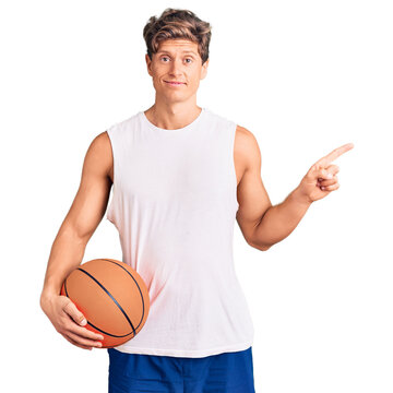 Young handsome man holding basketball ball smiling happy pointing with hand and finger to the side