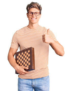 Young handsome man holding chess smiling happy and positive, thumb up doing excellent and approval sign