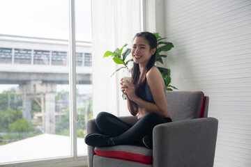 A Sporty young woman enjoy drinking proteins shake after doing morning exercise in living room, beautiful Asian female wearing sportswear doing weight training or practicing muscles and body workout. - obrazy, fototapety, plakaty