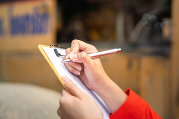 Action of an engineer hand is writing to perform inspection of the road roller or road compactor machinery engine part (as blurred background). Industrial working action scene, selective focus. 