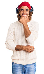 Young hispanic man listening to music using headphones looking confident at the camera smiling with crossed arms and hand raised on chin. thinking positive.