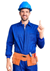 Young handsome man wearing worker uniform and hardhat with a big smile on face, pointing with hand finger to the side looking at the camera.