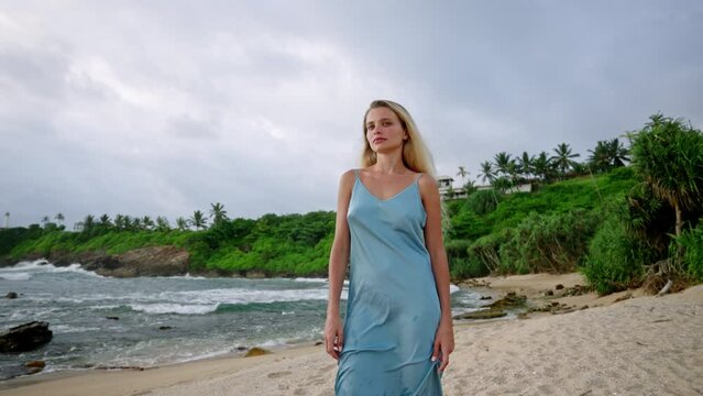 Bronzed woman in elegant blue gown strolls on sandy beach. Luxurious summer dress flows in breeze, high-end fashion shoot, perfumery ad appeal, exotic seascape backdrop, leisure tropical getaway.