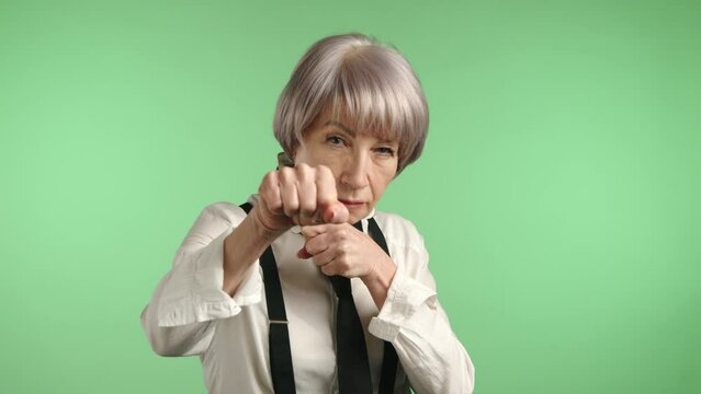 A determined senior woman demonstrating a self-defense stance with her fists ready, portraying strength and empowerment on a green background. Camera 8K RAW. 