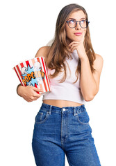 Beautiful caucasian young woman holding popcorn serious face thinking about question with hand on chin, thoughtful about confusing idea