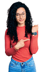 Young hispanic woman with curly hair wearing glasses in hurry pointing to watch time, impatience,...