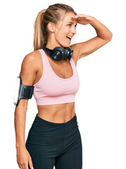 Young blonde woman wearing gym clothes and using headphones very happy and smiling looking far away with hand over head. searching concept.