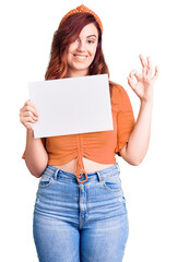 Young beautiful woman holding cardboard banner with blank space doing ok sign with fingers, smiling friendly gesturing excellent symbol