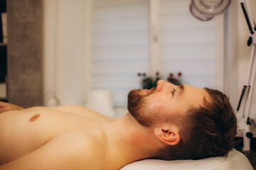 Beautician applying scrub onto young man's face in spa salon