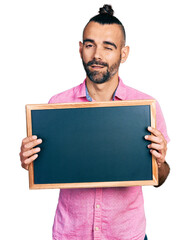 Hispanic man with ponytail holding blackboard winking looking at the camera with sexy expression,...