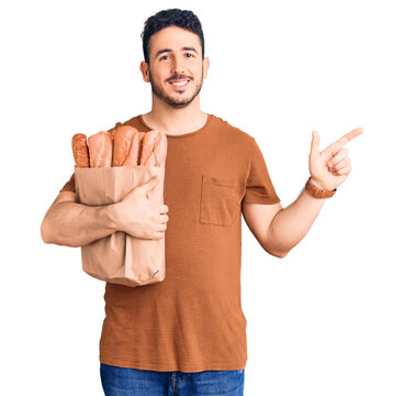Young hispanic man holding paper bag with bread smiling happy pointing with hand and finger to the side