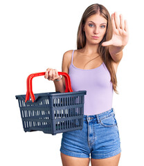 Young beautiful blonde woman holding supermarket shopping basket with open hand doing stop sign with serious and confident expression, defense gesture