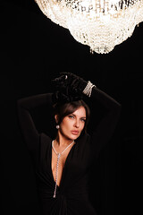 Beautiful woman in long black evening dress, celebrate New Year, Christmas Tree behind. Big lustre. Posch middle-aged lady with gorgeous jewelry of pearls. Portrait of girl in black room. 