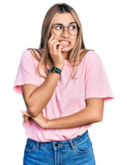 Hispanic young woman wearing casual t shirt and glasses looking stressed and nervous with hands on mouth biting nails. anxiety problem.