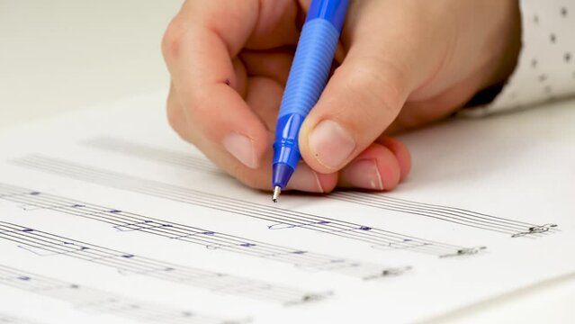 Closeup of the hand of a child holding a pen and writing on a piece of paper. An unknown child is studying in a music school. The child studies notes, music. A sheet music