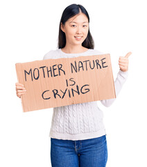 Young beautiful chinese woman holding mother nature is crying cardboard banner smiling happy and positive, thumb up doing excellent and approval sign