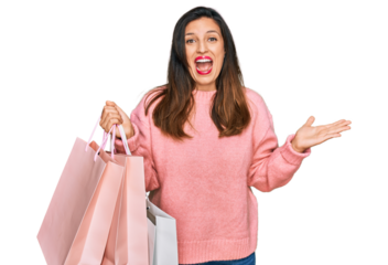 Beautiful hispanic woman holding shopping bags celebrating victory with happy smile and winner expression with raised hands