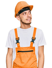 Hispanic young man wearing handyman uniform and safety hardhat looking away to side with smile on face, natural expression. laughing confident.