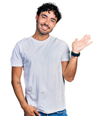 Hispanic young man with beard wearing casual white t shirt smiling cheerful presenting and pointing with palm of hand looking at the camera.