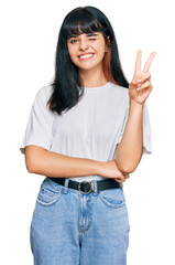 Young hispanic girl wearing casual clothes smiling with happy face winking at the camera doing victory sign. number two.