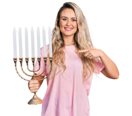 Young blonde woman holding menorah hanukkah jewish candle smiling happy pointing with hand and finger